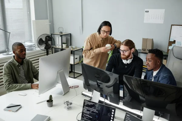 A diverse team of four IT professionals collaborating at a shared workstation in a modern office, reviewing code and data on multiple computer monitors, Veeam Backup Cloud Providers.